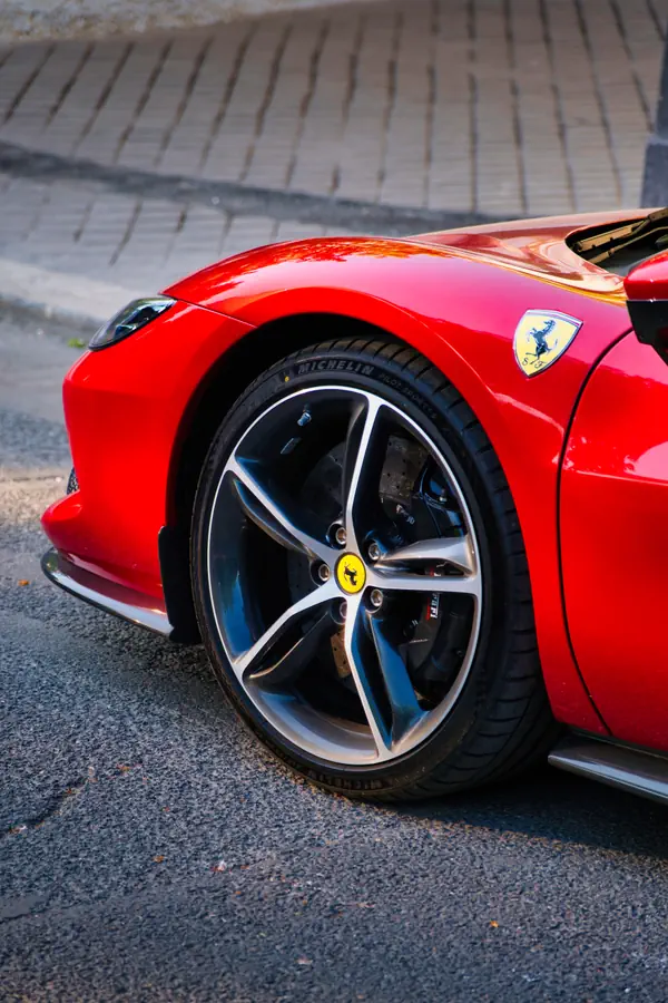 Gros plan sur la roue avant d'une Ferrari rouge, montrant la jante en alliage à cinq branches avec finition bicolore, l'étrier de frein, le pneu Michelin et l'emblème du cheval cabré sur l'aile.