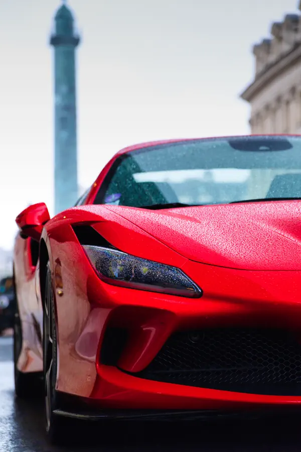 Gros plan sur l'avant d'une Ferrari F8 Tributo rouge sur la Place Vendôme à Paris, avec la colonne Vendôme visible en arrière-plan.