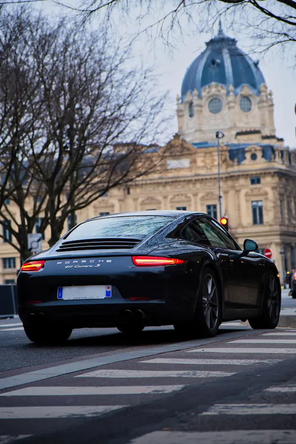 Porsche 911 Carrera S noire photographiée de trois-quarts arrière, avec ses feux LED caractéristiques allumés, devant un bâtiment parisien à dôme bleu ardoise