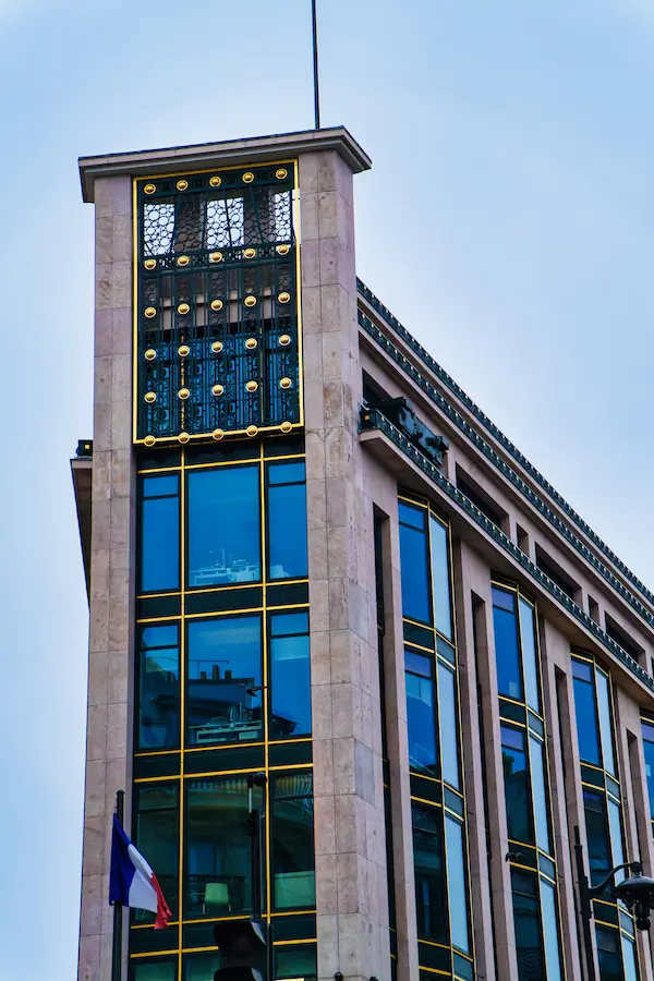 Angle d'un immeuble moderne parisien avec sa façade en pierre et verre, ornée de détails architecturaux dorés et d'une grille décorative art déco au sommet, avec un drapeau français en façade
