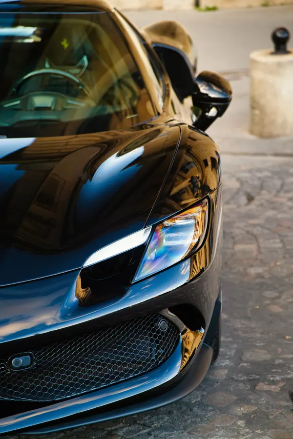 Gros plan sur l'avant d'une Ferrari 296 GTB noire, montrant le phare LED distinctif avec sa signature lumineuse caractéristique, la calandre en nid d'abeille et le capot sculpté. La voiture est stationnée sur des pavés parisiens.
