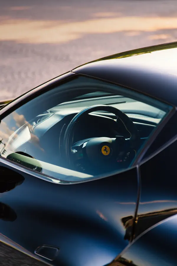 Vue à travers la vitre latérale d'une Ferrari noire, montrant le volant sportif avec l'emblème du cheval cabré au centre.