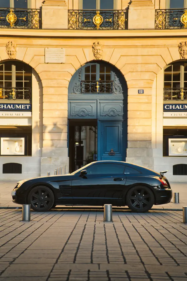Chrysler Crossfire noire personnalisée stationnée devant la boutique Chaumet sur la Place Vendôme à Paris. Cette voiture de sport américano-allemande au design distinctif est photographiée de profil, mettant en valeur sa silhouette allongée avec son long capot et son arrière tronqué.