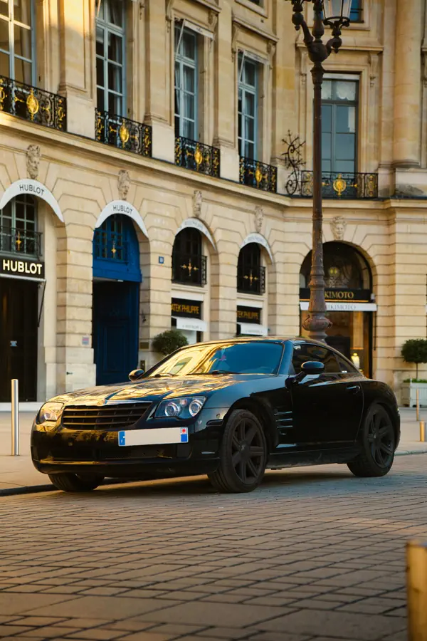 Chrysler Crossfire noire personnalisée avec jantes noires, photographiée sur la Place Vendôme à Paris au coucher du soleil. La voiture est stationnée devant les boutiques de luxe Hublot et Patek Philippe, dont les devantures sont visibles dans le bâtiment historique à la façade dorée par la lumière du soir.