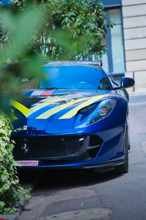 Ferrari 812 Superfast bleue foncée stationnée dans une rue urbaine, partiellement cachée par des feuillages verts. La voiture arbore des bandes décoratives jaunes et bleues sur le capot ainsi que le célèbre emblème du cheval cabré.