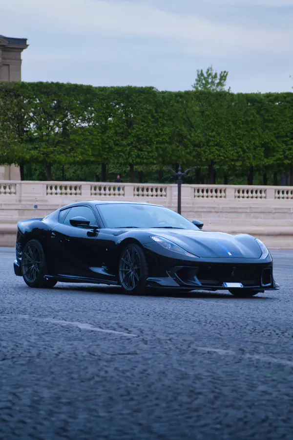 Ferrari 812 Competizione noire photographiée sur une large avenue parisienne, avec sa silhouette élancée et ses lignes aérodynamiques caractéristiques. En arrière-plan, une balustrade en pierre blanche et une rangée d'arbres verdoyants d'un jardin public parisien sous un ciel légèrement nuageux.