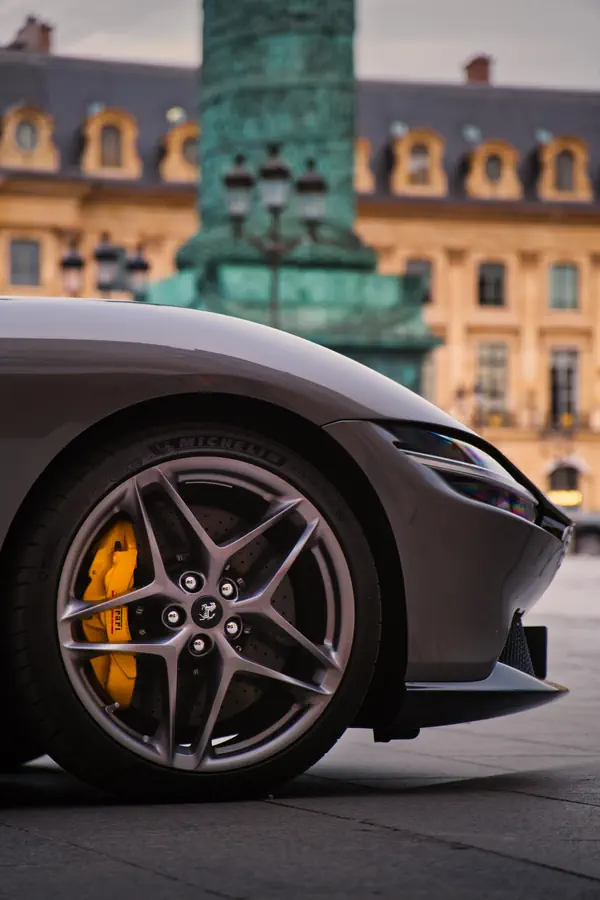 Gros plan sur la roue avant d'une Ferrari Roma gris métallisé sur la Place Vendôme, montrant la jante en alliage à cinq branches, l'étrier de frein jaune Ferrari et le pneu Michelin. En arrière-plan, on distingue la colonne Vendôme en bronze vert-de-gris et les façades dorées des bâtiments historiques de la place.