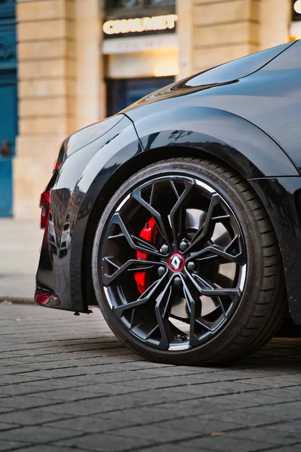 Gros plan sur la roue arrière d'une Renault Megane RS noire avec jantes en alliage noir géométriques, étriers de frein rouges visibles, garée sur une rue pavée devant un bâtiment historique en pierre.