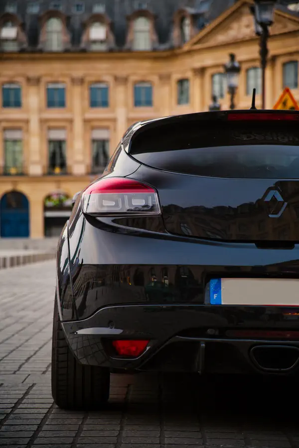 Arrière d'une Renault Megane RS noire garée sur une place pavée. En arrière-plan, un élégant bâtiment historique parisien aux façades beiges et toiture mansardée.