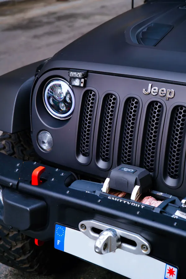 Gros plan sur l'avant d'un Jeep Wrangler noir mat équipé d'un treuil Smittybilt et de phares LED circulaires. La calandre à sept fentes caractéristique arbore le logo Jeep chromé.