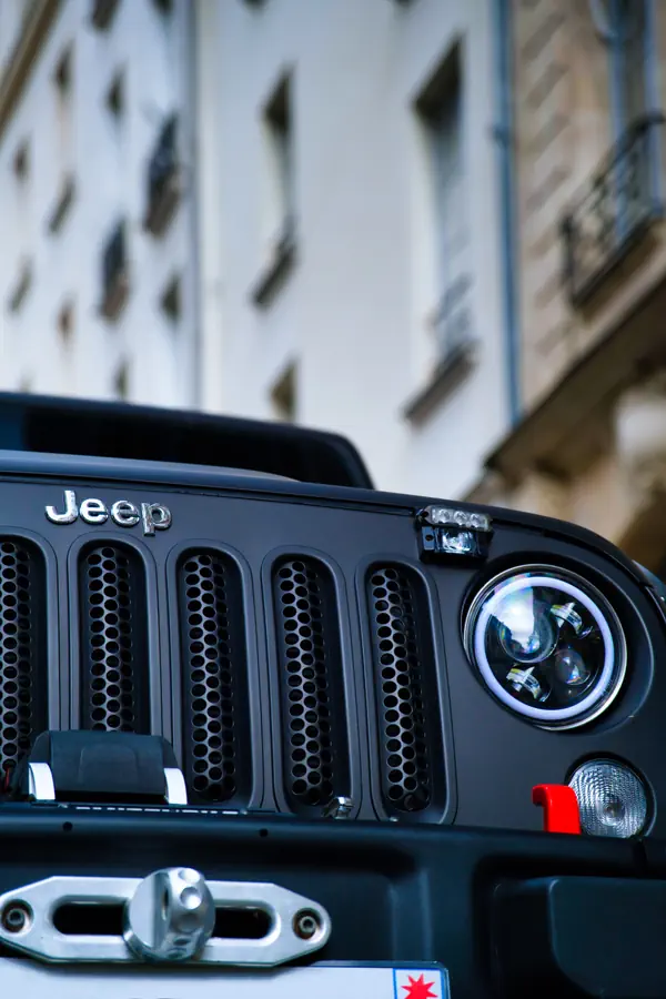 Gros plan sur la calandre à sept fentes d'un Jeep Wrangler noir mat avec logo Jeep chromé, phare LED rond avec halo lumineux, barre LED auxiliaire sur le capot et crochet de remorquage rouge. En arrière-plan flou, une façade d'immeuble parisien traditionnel.