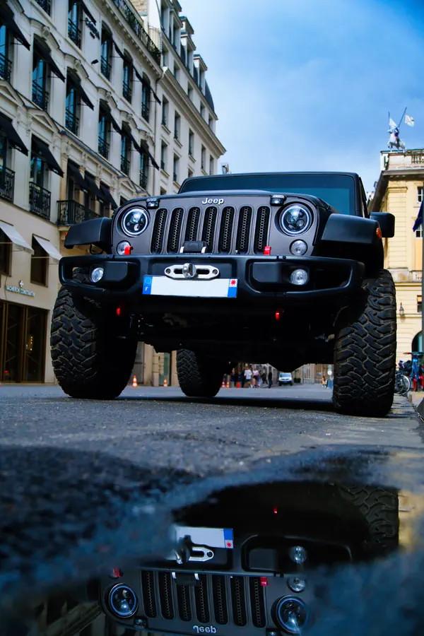 Vue frontale d'un Jeep Wrangler noir mat surélevé avec pneus tout-terrain, photographié au ras du sol avec reflet dans une flaque d'eau. La scène se déroule dans une rue parisienne élégante bordée d'immeubles haussmanniens, avec une statue visible au loin sur un toit.