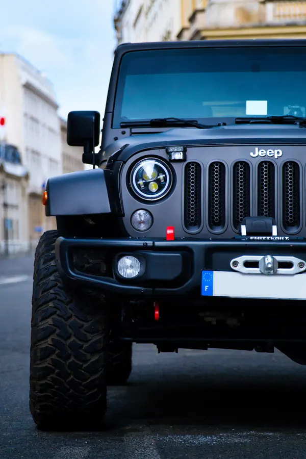 Vue de face d'un Jeep Wrangler noir mat avec phares LED ronds, calandre à sept fentes, treuil Smittybilt et pneus tout-terrain à motif agressif. Le véhicule est stationné dans une rue parisienne avec des immeubles historiques en arrière-plan.