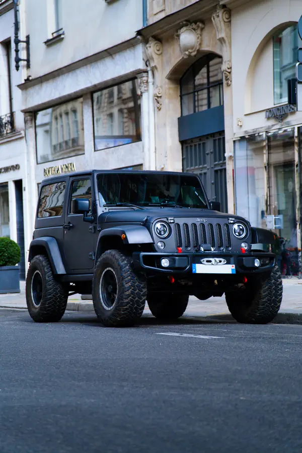 Jeep Wrangler noir mat à deux portes stationné dans une rue parisienne élégante et avec des pneus tout-terrain surdimensionnés. En arrière-plan, des façades d'immeubles haussmanniens abritant des boutiques de luxe, dont Bottega Veneta.