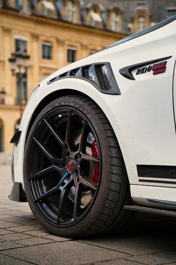 Détail de l'aile avant d'une Mercedes-Benz E63 S AMG Brabus blanche avec l'insigne "RENNTECH 800" et une prise d'air latérale en fibre de carbone. La roue présente une jante noire au design sportif avec étriers de frein rouges visibles.