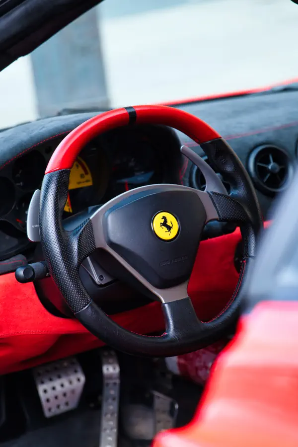 Intérieur d'une Ferrari avec volant en cuir noir perforé et bande rouge au sommet, orné du célèbre emblème du cheval cabré au centre.