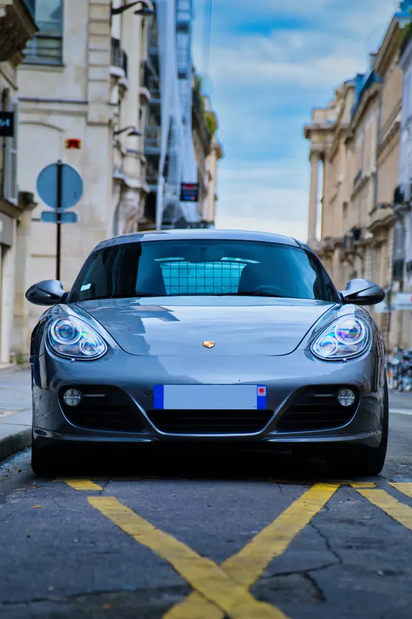 Porsche Cayman gris métallisé photographié de face, stationné dans une rue parisienne avec marquage au sol jaune.