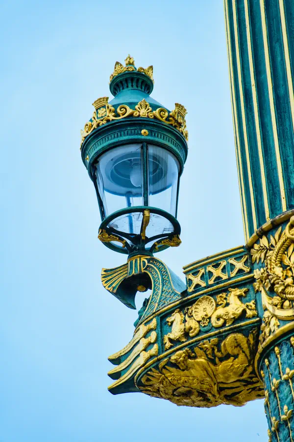 Détail d'un lampadaire historique parisien, avec sa lanterne couronnée d'ornements dorés et sa console richement décorée de motifs végétaux et architecturaux en vert et or, typique du style Second Empire
