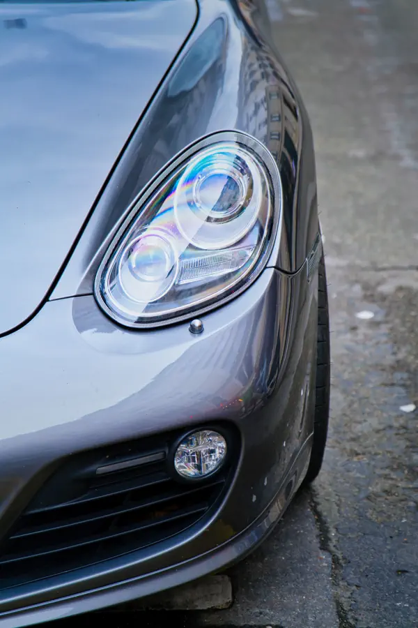 Gros plan sur le phare avant d'une Porsche Cayman gris métallisé, montrant le design distinctif bi-xénon avec anneau LED caractéristique et antibrouillard intégré dans le pare-chocs inférieur.