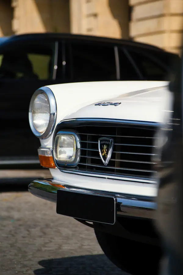 Avant d'une Peugeot 404 blanche classique, montrant sa calandre chromée horizontale avec l'emblématique écusson Peugeot au centre, ses phares ronds caractéristiques et ses clignotants rectangulaires.