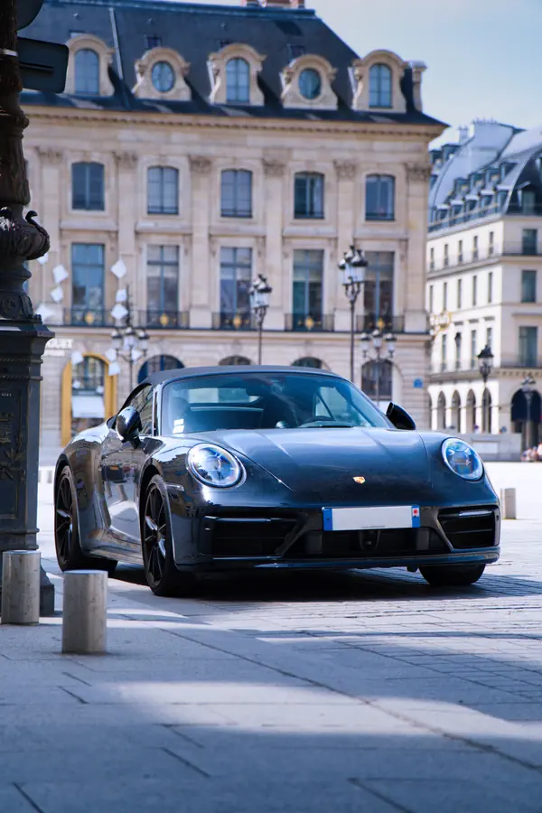 Porsche 911 noire (modèle récent) photographiée sur la Place Vendôme à Paris, avec ses phares LED ronds caractéristiques et sa silhouette emblématique. En arrière-plan, on distingue les façades élégantes des bâtiments historiques de la place avec leurs fenêtres mansardées, leurs lampadaires ornés et leurs arcades.