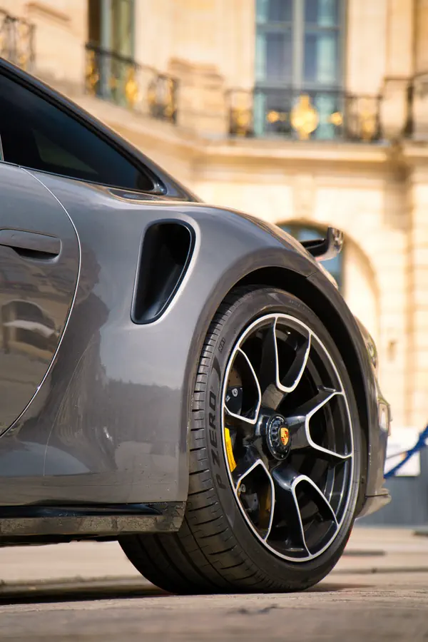 Gros plan sur la roue arrière d'une Porsche 911 Turbo gris métallisé, montrant sa jante noire au design complexe avec finition diamantée, les étriers de frein jaunes et la prise d'air latérale caractéristique du modèle Turbo.