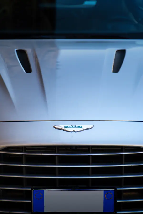 Gros plan sur l'avant d'une Aston Martin blanche, montrant l'emblématique badge ailé de la marque britannique au-dessus de la calandre horizontale caractéristique.