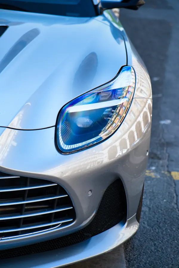 Gros plan sur le phare avant d'une Aston Martin blanche, montrant son design élégant avec bande LED distinctive et finition cristalline.