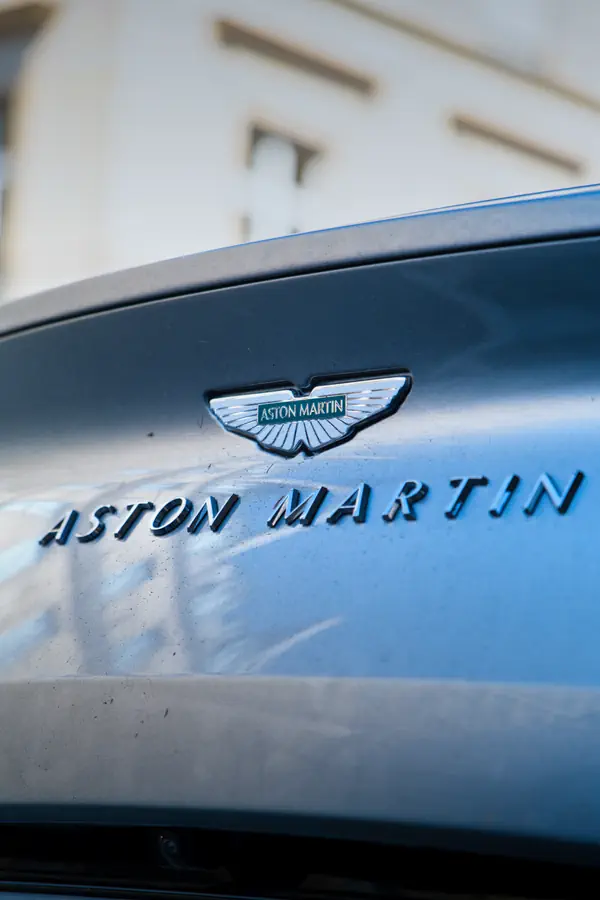 Gros plan sur le badge emblématique Aston Martin - ailes stylisées sur fond vert - et le lettrage chromé "ASTON MARTIN" apposés sur la carrosserie gris métallisé d'une voiture de la marque britannique.