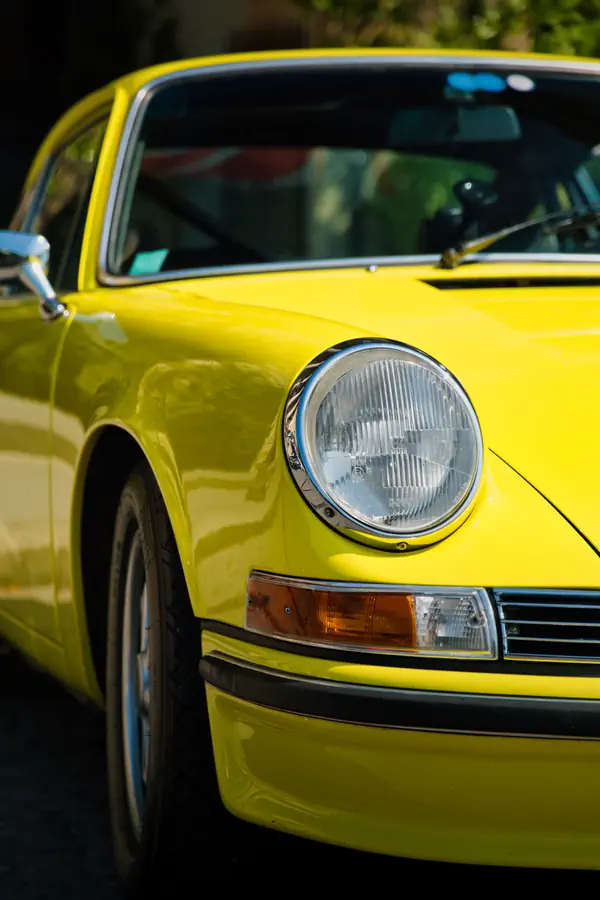 Gros plan sur l'avant d'une Porsche 911 classique jaune vif, mettant en évidence son phare rond caractéristique à cerclage chromé et son clignotant orange rectangulaire.