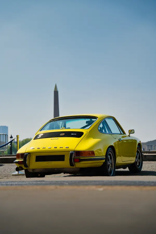 Porsche 911 classique jaune photographiée de trois-quarts arrière sur une esplanade parisienne, avec l'obélisque de la Place de la Concorde visible en arrière-plan.