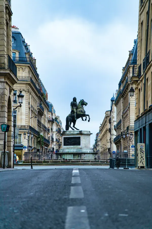 Statue équestre encadrée par des immeubles haussmanniens aux toits en ardoise bleue, vue depuis le sol avec une perspective marquée par les lignes blanches de la chaussée, créant une composition symétrique typiquement parisienne