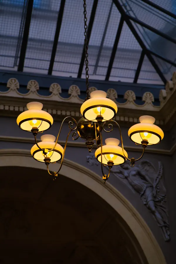 Lustre art nouveau à cinq globes lumineux en verre opalin blanc, aux bras courbes en bronze, suspendu sous une verrière ornée de moulures décoratives, créant une atmosphère chaleureuse avec sa lumière dorée