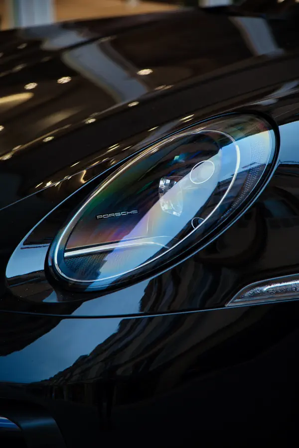 Gros plan sur le phare avant d'une Porsche noire, montrant le design sophistiqué de l'optique LED avec sa signature lumineuse caractéristique et le logo Porsche intégré, reflétant la lumière sur sa carrosserie brillante