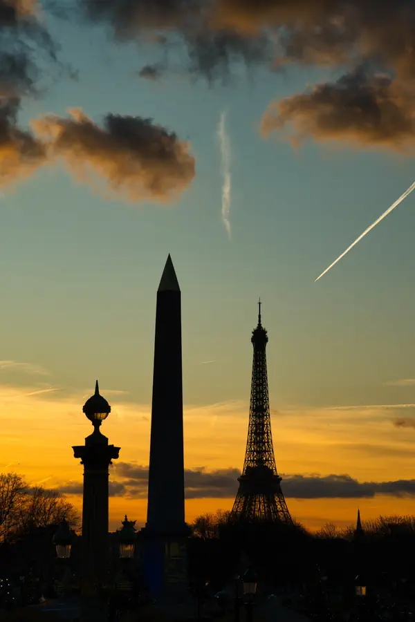 Silhouettes de l'obélisque de la Concorde, d'un lampadaire historique et de la Tour Eiffel se détachant sur un ciel crépusculaire aux teintes orangées, avec des traînées d'avion traversant le ciel