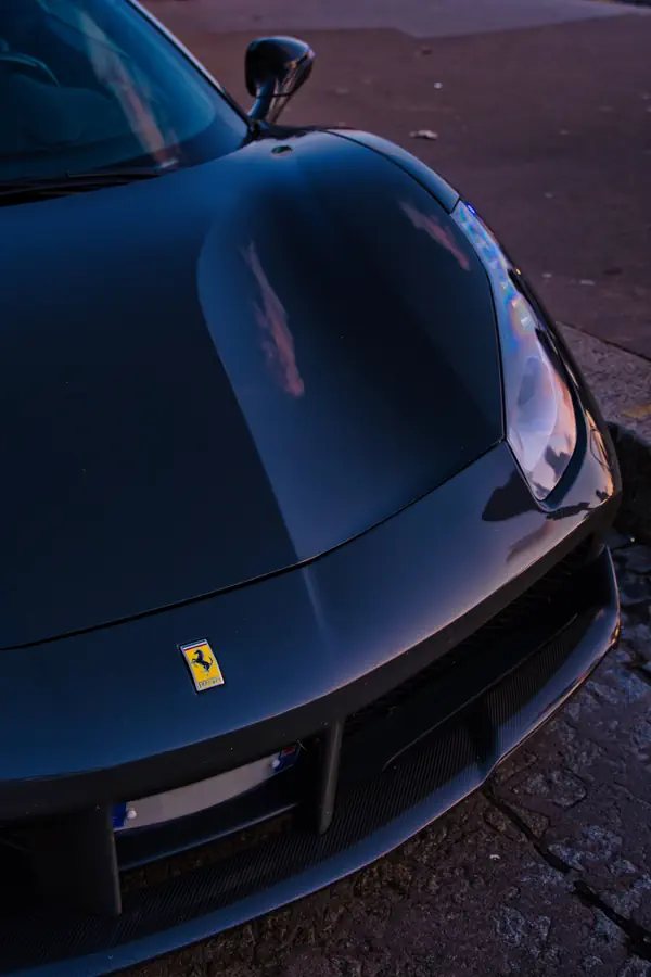 Détail du capot avant d'une Ferrari noire, mettant en valeur l'emblématique écusson au cheval cabré et les lignes aérodynamiques de la carrosserie, photographié au crépuscule