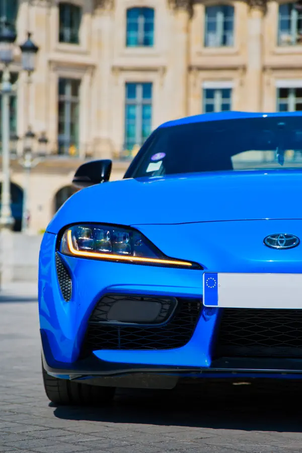 Avant d'une Toyota Supra bleue électrique avec phares LED distinctifs et calandre noire sportive, stationnée devant une façade classique de couleur crème.