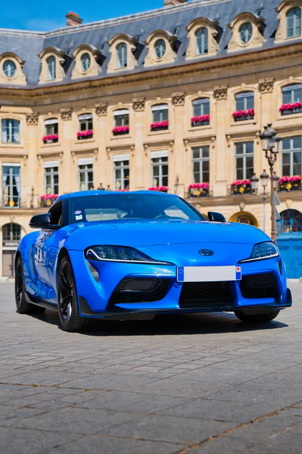 Toyota Supra bleue électrique photographiée de face sur une place pavée, devant un élégant bâtiment historique français de style haussmannien avec toit mansardé en ardoise et façade en pierre beige ornée de jardinières de fleurs roses.
