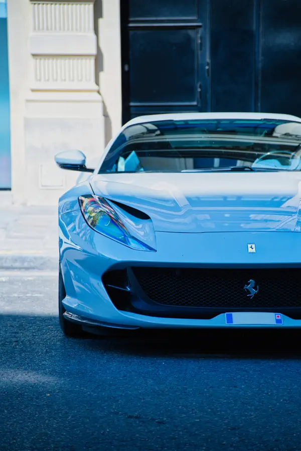 Vue frontale rapprochée d'une Ferrari 812 Superfast stationnée dans une rue urbaine. L'image met en valeur le phare effilé, la calandre noire avec l'emblème du cheval cabré, et les lignes aérodynamiques du capot.