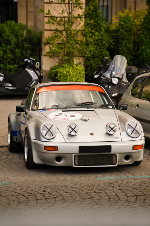 Porsche 911 de rallye blanche avec bande orange sur le pare-brise, arborant le numéro 259 sur un médaillon rond. Les quatre phares sont protégés par des croix en ruban adhésif, caractéristique des voitures de compétition.