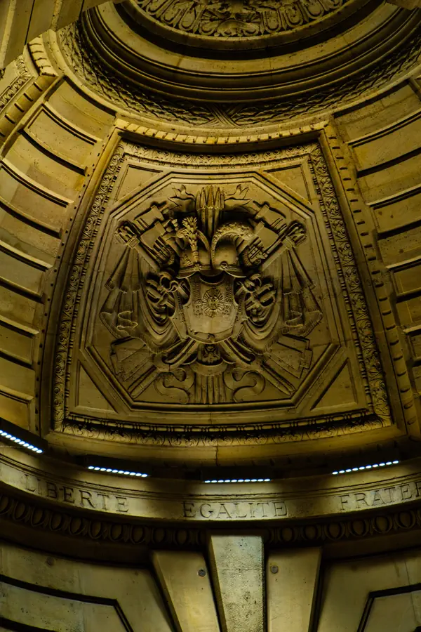 Détail architectural d'un bâtiment officiel parisien montrant un blason sculpté en relief avec des symboles républicains, surmonté d'une couronne, et la devise "LIBERTE EGALITE FRATERNITE" gravée en dessous, éclairé dans des tons dorés