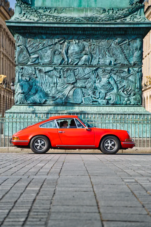 Porsche 911 classique rouge photographiée de profil sur une place pavée, devant le socle en bronze patiné d'un monument historique orné de bas-reliefs détaillés.