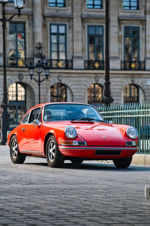 Porsche 911 classique rouge vif photographiée sur une place pavée devant un élégant bâtiment historique parisien avec façade en pierre, balcons en fer forgé ornés de dorures et lampadaires d'époque.