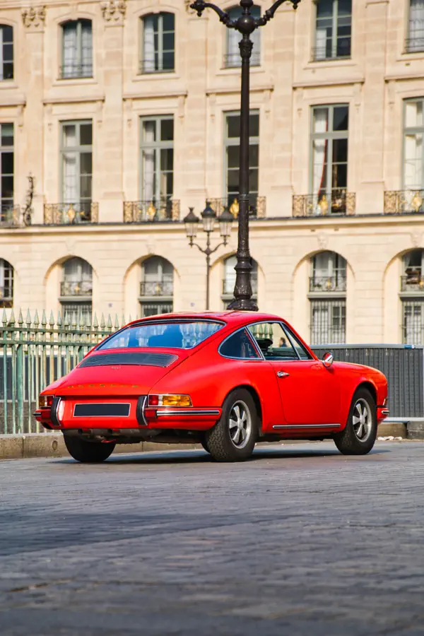 Vue arrière trois-quarts d'une Porsche 911 classique rouge stationnée sur une place pavée parisienne.