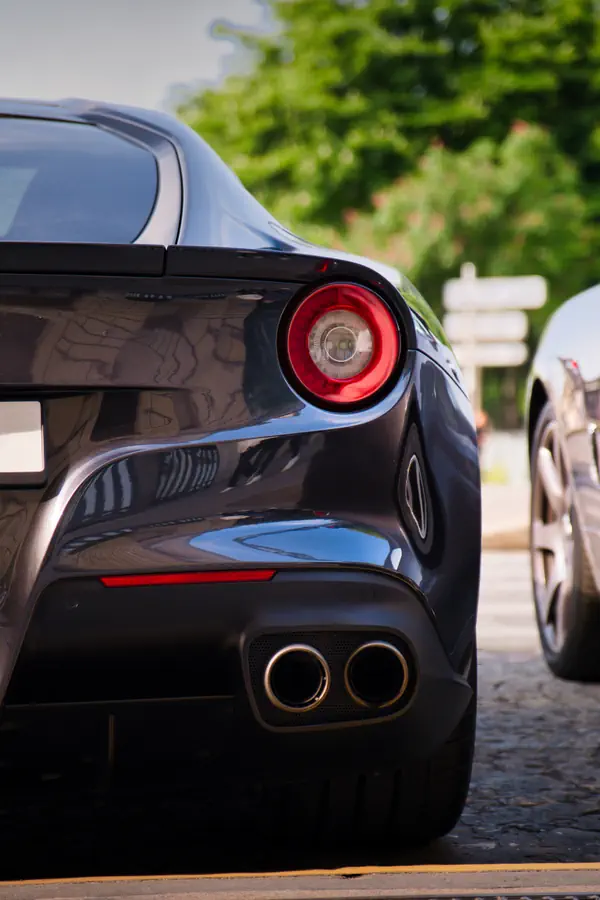 Détail de l'arrière d'une Ferrari F12 Berlinetta noire montrant son feu rond caractéristique avec anneau lumineux rouge, ses doubles sorties d'échappement chromées et ses lignes aérodynamiques sculptées.