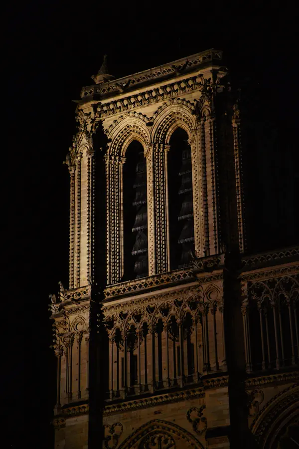 Détail architectural de Notre-Dame de Paris photographié de nuit, montrant ses fenêtres gothiques en ogive, ses colonnes ornées et sa galerie sculptée, illuminées dans des tons dorés qui contrastent avec l'obscurité environnante