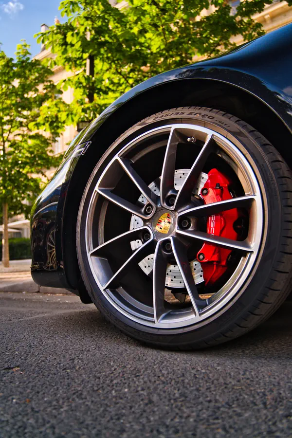 Gros plan sur la roue avant d'une Porsche noire montrant une jante en alliage gris bicolore à dix branches avec l'écusson Porsche au centre, un étrier de frein rouge et un disque de frein perforé.