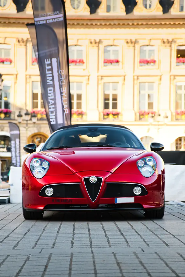 Alfa Romeo 8C Spider rouge photographiée de face sur une place pavée, devant un élégant bâtiment historique avec façade jaune ornée de jardinières de fleurs roses.