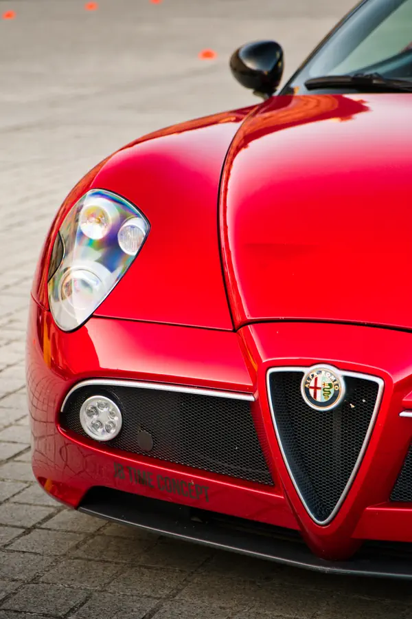 Gros plan sur l'avant d'une Alfa Romeo 8C Spider rouge vif, montrant son phare triple distinctif, sa calandre triangulaire emblématique avec l'écusson Alfa Romeo, et son antibrouillard LED rond intégré dans la grille noire.