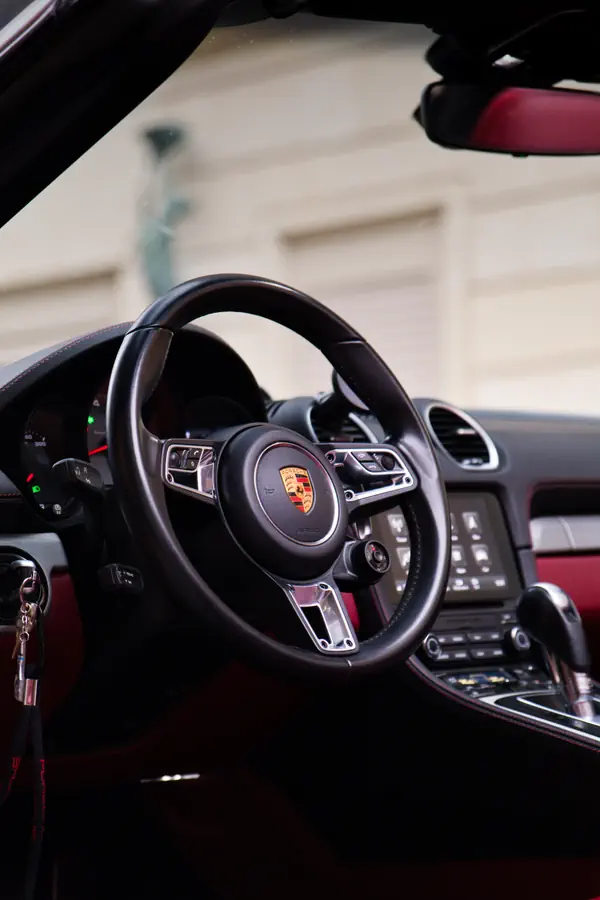 Intérieur de Porsche avec volant sport en cuir noir orné du logo Porsche, tableau de bord moderne avec écran tactile central et sellerie rouge et noire.